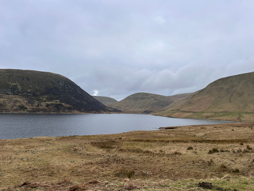 Serene landscape featuring a calm lake nestled between gently sloping hills. The hills are predominantly brown and green, indicating a likely temperate climate. The sky is overcast, casting a soft, diffused light across the scene. The scene suggests a remote and sparsely populated area.