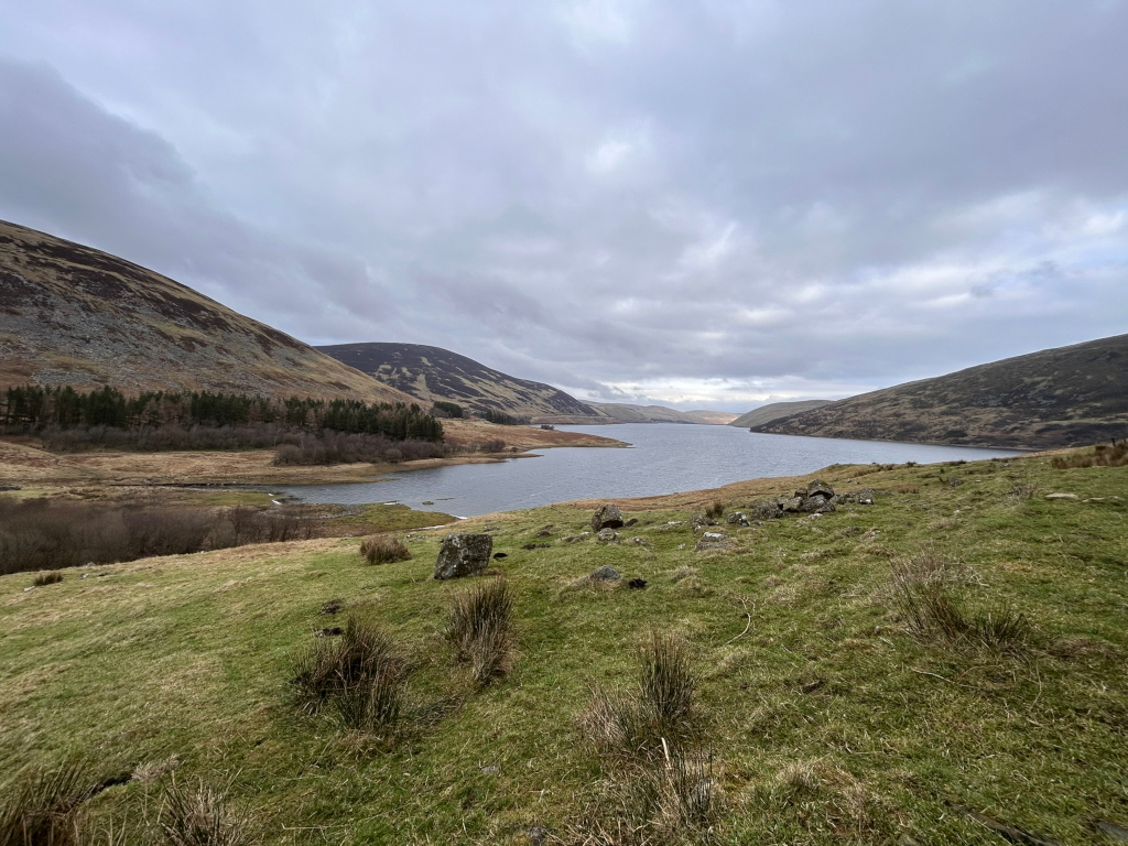 Serene landscape featuring a calm body of water nestled between rolling hills under a cloudy sky. The foreground is composed of grassy hills, speckled with rocks. A small cluster of rocks is visible in the mid-ground, near the water's edge. In the background, the hills are covered in sparse vegetation, and a line of trees can be seen on the left. The overall atmosphere is peaceful and somewhat muted in colour due to the overcast sky.