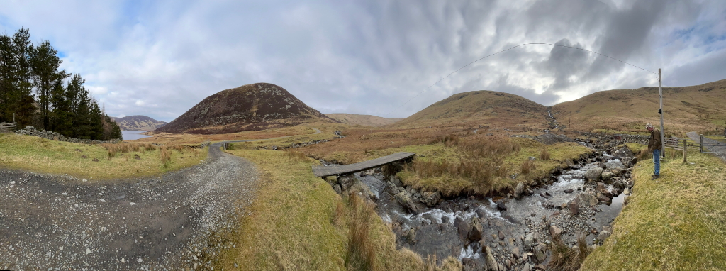 Panoramic view of a rural landscape featuring a small wooden bridge spanning a rocky stream, which flows towards the viewer. A dirt road forks in the foreground, with one branch leading towards the stream and the other turning away. In the background are rolling hills, one of them a distinct, dome-shaped hill. Charlie stands near the stream on the right side of the image, seemingly observing the scene. The sky is cloudy. The overall impression is of a serene and possibly remote location.
