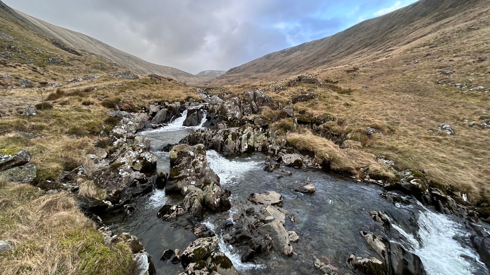 Mountain stream cascading down a rocky ravine. The stream is relatively narrow, with the water flowing over dark rocks and appearing clear and fast-moving. The surrounding landscape is characterised by dry, brownish-gold grasses covering the gently sloping hillsides under a somewhat cloudy sky. The overall impression is one of a remote, wild, and possibly somewhat bleak, but also beautiful, natural setting.