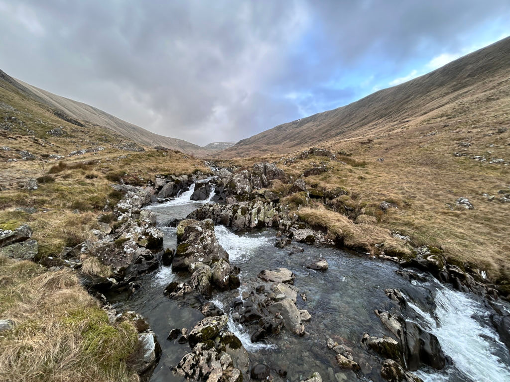 Mountain stream cascading down a rocky ravine. The stream is relatively narrow, with the water flowing over dark rocks and appearing clear and fast-moving. The surrounding landscape is characterised by dry, brownish-gold grasses covering the gently sloping hillsides under a somewhat cloudy sky. The overall impression is one of a remote, wild, and possibly somewhat bleak, but also beautiful, natural setting.