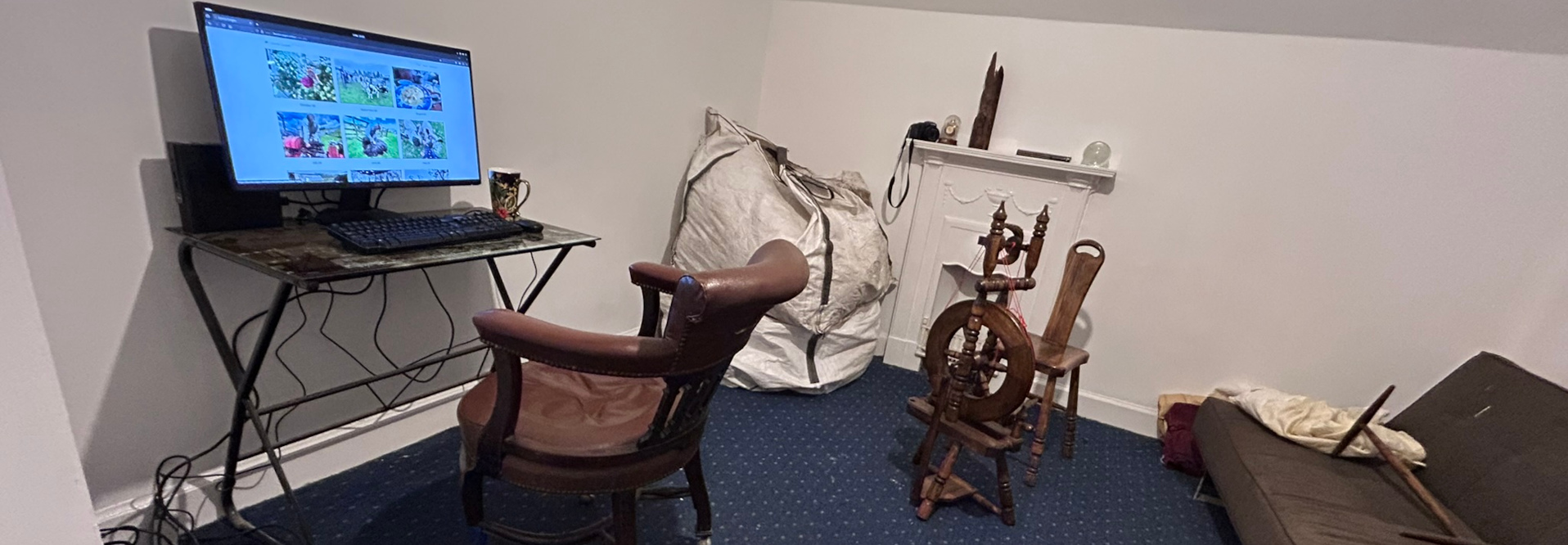 A corner of a room with a computer setup on a small metal desk. A brown leather chair sits in front of the desk. A large white burlap bag is partially visible near the desk. In the corner, there's an antique spinning wheel and a small, dark-coloured couch with a light-coloured cloth draped on it. A white mantelpiece is on the wall.