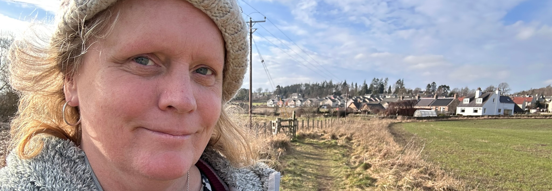 Close-up of a fair-skinned woman with blonde hair, wearing a grey knitted hat and a grey fluffy jacket. She, Leonie, is smiling slightly and looking directly at the camera. The background is blurred but shows a rural landscape with a small village in the distance under a partly cloudy blue sky. A path or track runs from the foreground towards the village.