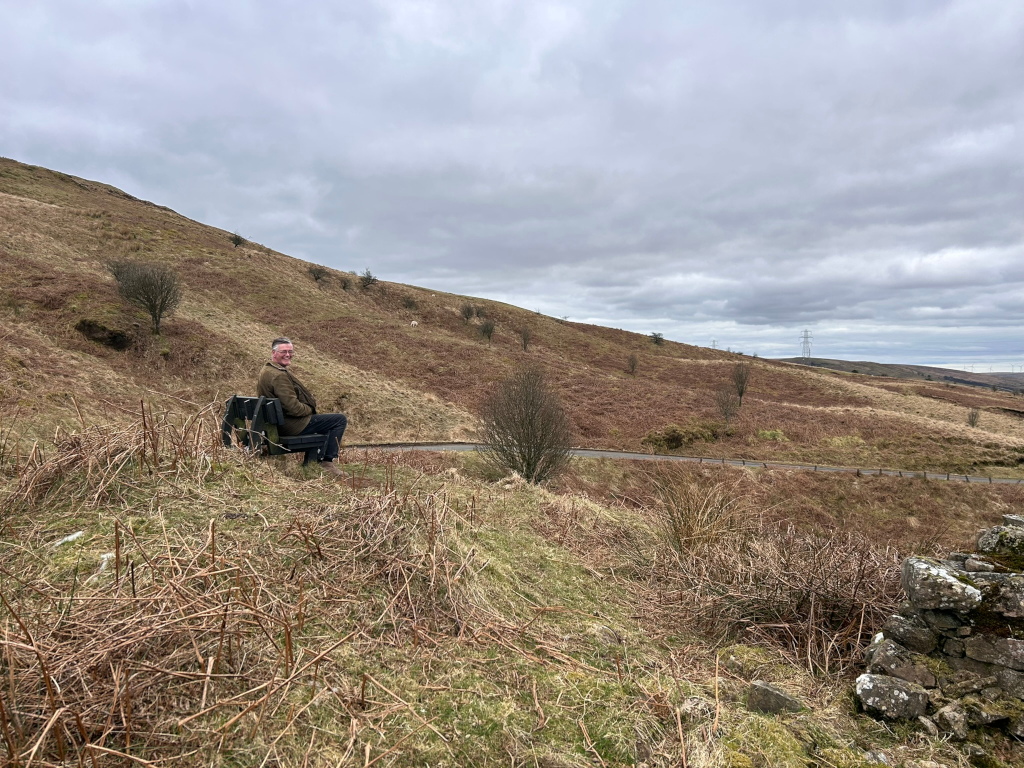 Charlie sitting on a park bench on a hillside. The hillside is sparsely vegetated with brown grasses and shrubs. A road is visible in the mid-ground, and in the far background, there is a more distant hill and a power line. The sky is overcast. The overall mood is peaceful and somewhat desolate.