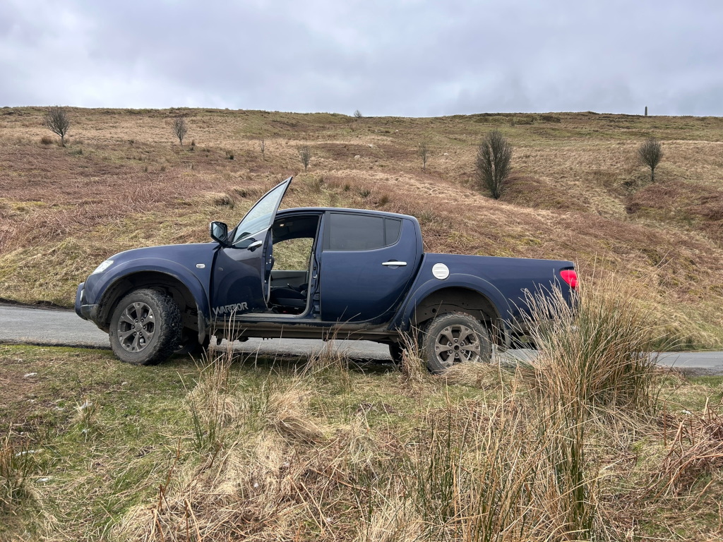 Dark blue Mitsubishi L200 Warrior pickup truck parked on the side of a road next to a grassy hill. The front passenger side door is open. The setting appears to be a rural or mountainous area, with dry, brown grass covering the hillside. The sky is overcast. The overall impression is one of a vehicle stopped during a journey in a remote, natural landscape.