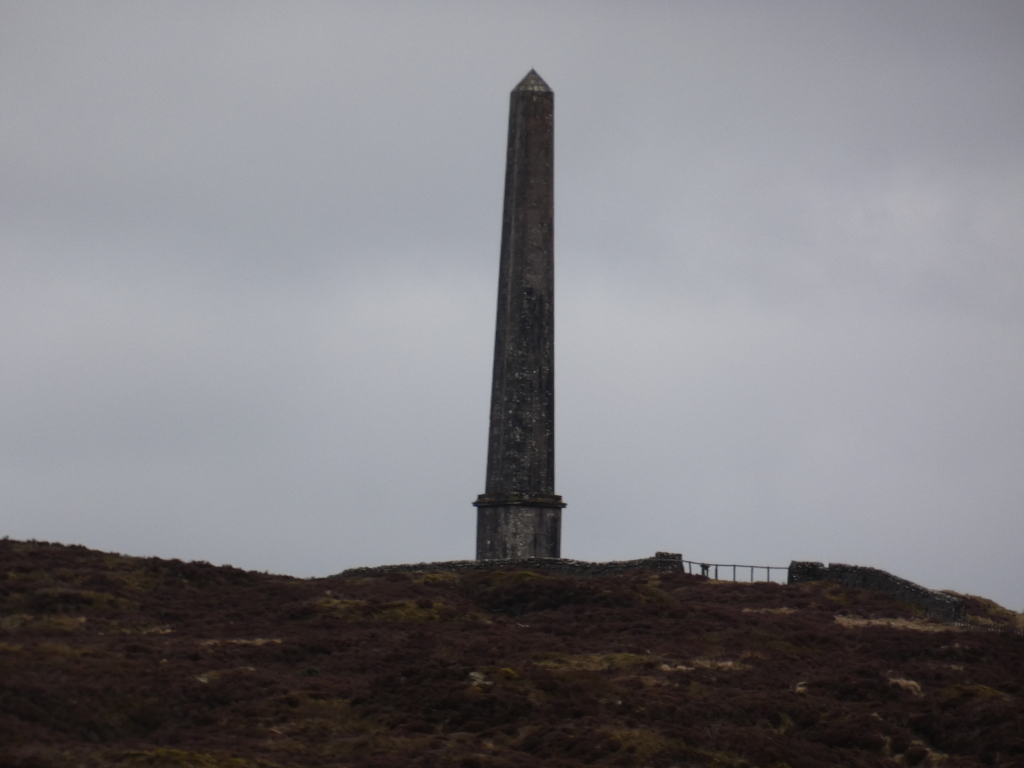 Tall, slender obelisk standing atop a heather-covered hill against a muted, overcast sky. The obelisk appears to be made of dark stone and is fairly weathered. A low stone wall partially encircles the base of the obelisk. The overall mood is sombre and somewhat desolate.