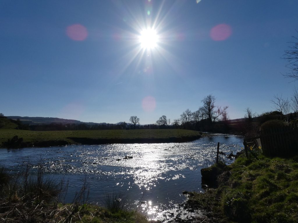 Tranquil river scene on a bright, sunny day. The sun is positioned high in the sky, its light creating sparkles on the water's surface. The river gently meanders through a grassy field, bordered by a few bare trees and a wooden fence. The background features rolling hills under a clear blue sky. The overall mood is peaceful and serene.