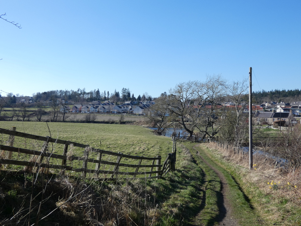 Dirt path leading towards a row of houses situated beyond a field. A wooden fence lines the path, and trees frame the scene, with a small stream or river visible in the middle ground. The sky is clear and blue, indicative of a sunny day. The overall impression is one of a rural or semi-rural setting, possibly in a village or small town.