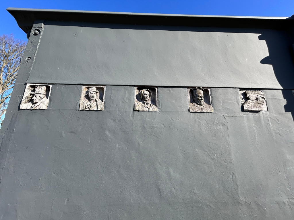 Dark grey wall with five recessed panels, each containing a bas-relief sculpted head. The heads appear to represent historical figures, possibly royalty or important people from a particular era or location, given the variety of headdresses and clothing styles. The style of the sculptures suggests a classical or historical representation. The overall effect is one of commemoration or homage.