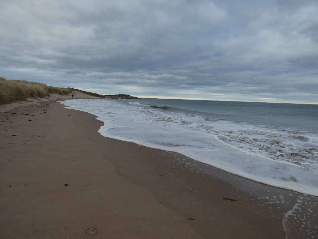 Beach scene under a cloudy sky. The sand is light brown, and waves are gently lapping the shore. A lone figure is visible walking along the beach in the distance. The overall mood is calm and somewhat melancholic.