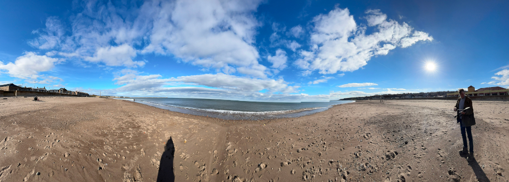 Panoramic view of a sandy beach on a sunny day. The beach stretches from the foreground to the calm sea in the distance. Charlie stands near the right side of the image, appearing to be holding a camera stand. Buildings and a seawall are visible in the background along the left and right sides of the beach. The sky is mostly clear with scattered, fluffy white clouds beneath a bright blue expanse. The overall impression is one of serenity and calm.