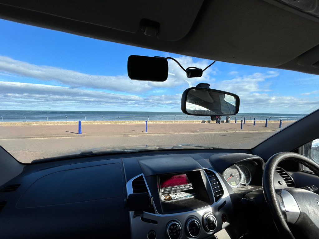The interior of a car parked near a seaside. The car's wind-shield provides a view of a calm ocean under a mostly sunny sky with some clouds.  Blue bollards are visible along the seawall. The photo appears to be taken on a bright, clear day. The focus is on the view outside the car, with the interior dashboard slightly out of focus.
