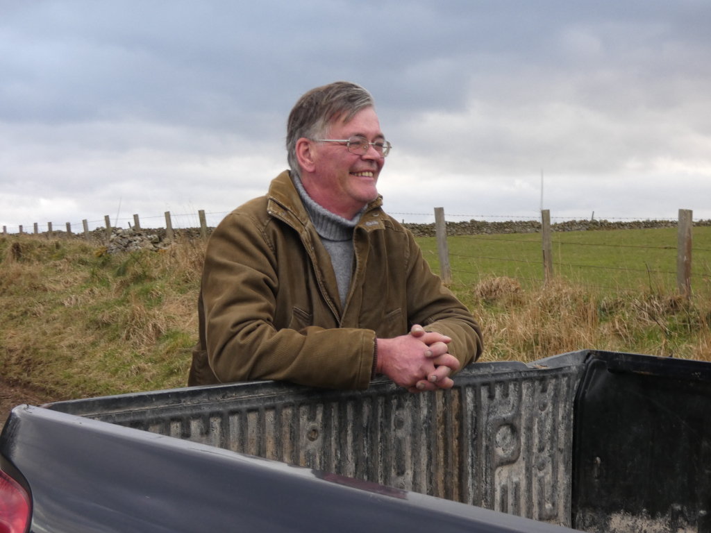 Charlie wearing glasses, smiling slightly, leaning against the back of a dark-coloured pickup truck. He's wearing a brown jacket and a dark-coloured turtle-neck sweater. The background features a rural landscape with a stone wall, a fence, and a grassy field under a cloudy sky. The overall impression is one of peaceful contemplation in a rural setting.