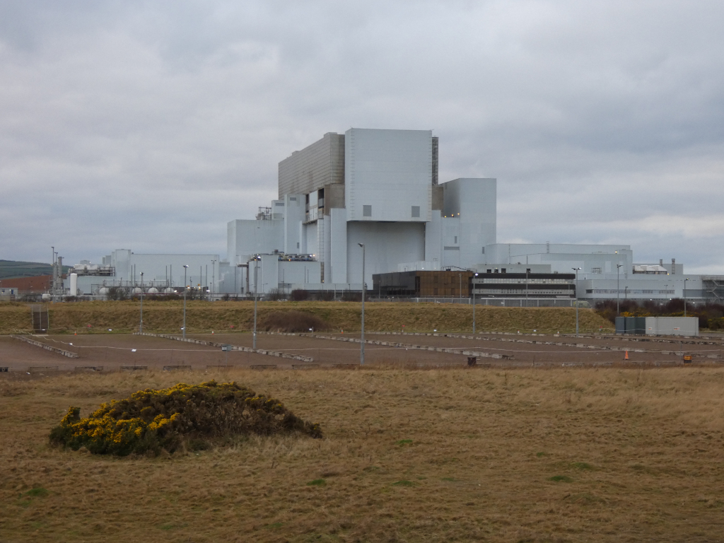 A large industrial facility, possibly a Nuclear power plant, situated in a relatively flat landscape. The facility is primarily white or light-coloured, with several buildings of varying heights. In the foreground, there's a brown, grassy field with a small bush, and what appears to be a large, empty parking lot or storage area in front of the facility. The sky is overcast and gray. The overall impression is one of industrial scale and a somewhat bleak, though not necessarily unpleasant, atmosphere.