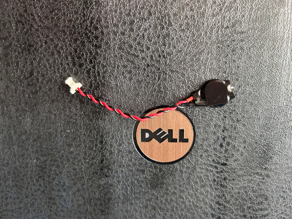 Small, round, black battery with a red and black wire attached. The wire connects to a small white connector. The battery is positioned near a circular, copper-coloured Dell logo on a dark textured surface. The overall impression is of a close-up shot of a Dell computer component, likely a CMOS battery.