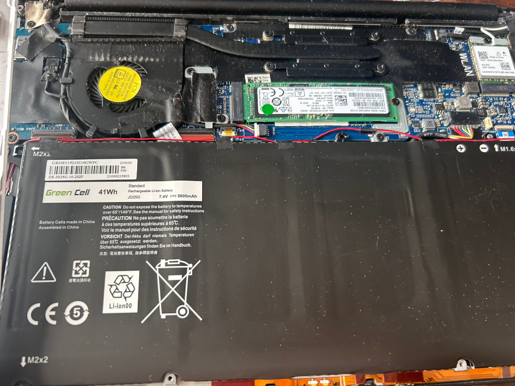 Internal components of a laptop, specifically focusing on the battery. The battery is a Green Cell 41Wh rechargeable Li-ion battery with various safety warnings and specifications printed on its surface. The image also reveals other components such as the cooling fan, a solid-state drive (SSD), and various circuit boards. The overall impression is a technical view of the laptop's internal workings, possibly taken during repair or maintenance. There is no artistic or emotional expression; the image is purely descriptive of a technological subject. There is no explicit story or moral conveyed.