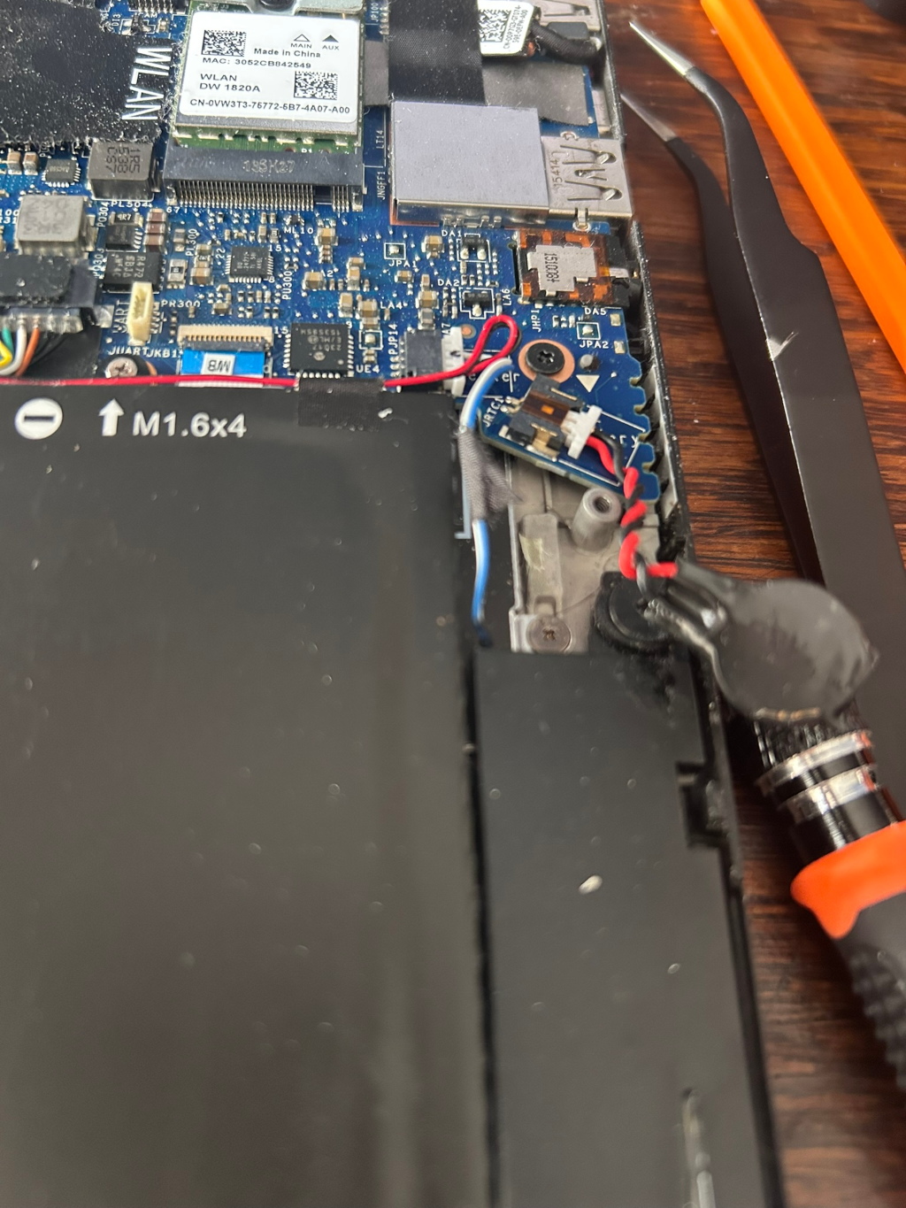 A close-up view of the interior of a laptop, specifically the motherboard and battery area during a repair or maintenance process. Various components are visible, including the motherboard with integrated circuits, connectors, and wires. A partially removed battery is shown, labelled M1.6x4, indicating a screw specification. Tweezers and a screwdriver are present, suggesting work in progress. The overall impression is a technical image capturing a step in a laptop repair.