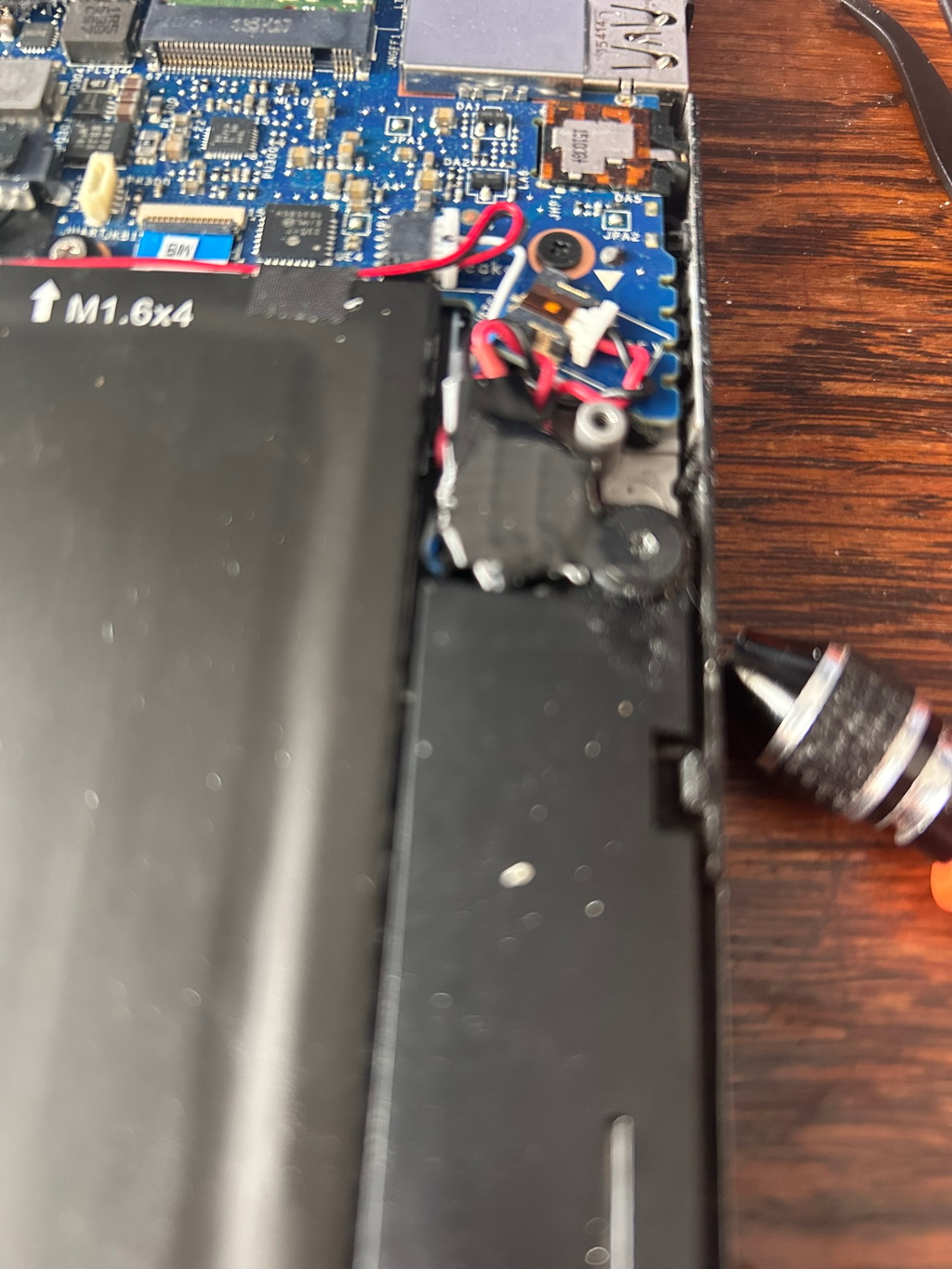 Inside of a disassembled laptop. The focus is on the motherboard and battery area, with various components visible, including wires, screws, and a small electronic part partially removed. The overall impression is of a laptop undergoing repair or maintenance. A small screwdriver is visible in the bottom right corner.