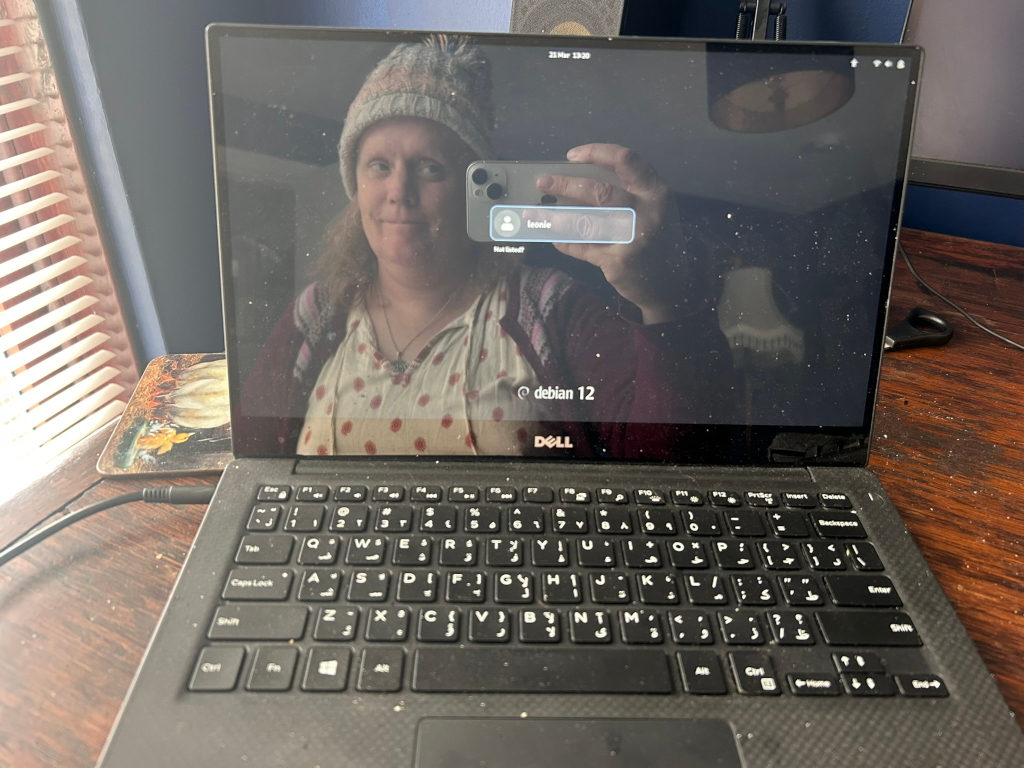 Dell laptop displaying a login screen. The screen also reflects the image of the user, a woman wearing a knit hat, taking a selfie with her phone to log in. The background is a dimly lit room, and the laptop itself appears to be dusty. The operating system shown is Debian 12. The overall impression is one of casual, everyday technology use. There is no deeper narrative or moral implied; it's a simple snapshot of an individual using their laptop.