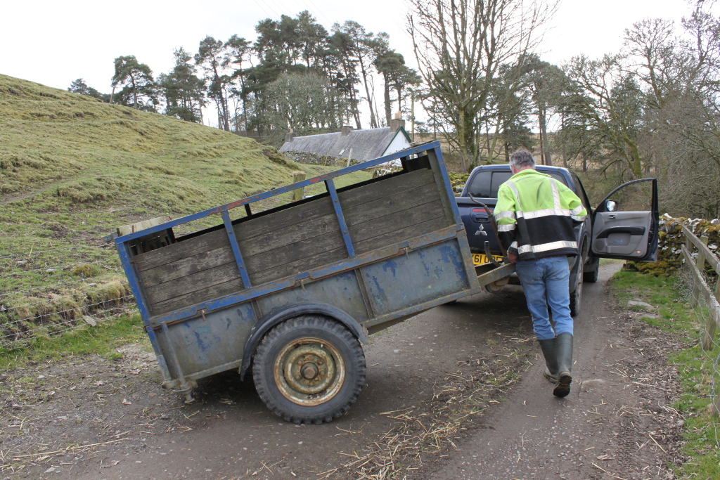 Charlie in a high-visibility jacket and Wellington boots manoeuvring a weathered, blue livestock trailer away from a dark-coloured pickup truck parked on a rural, unpaved road. The background includes a hillside, trees, and a small stone cottage. The overall impression is one of rural work or farm activity.