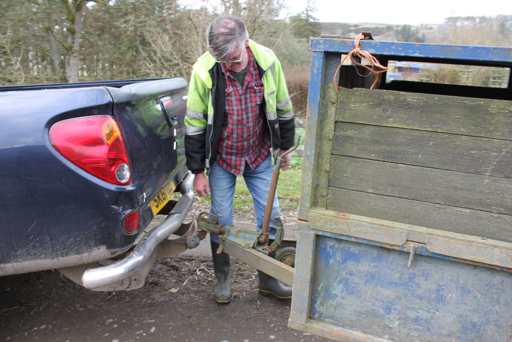 Charlie in work boots and a high-visibility jacket attaching a rusty, old trailer to the tow hitch of a dark-blue pickup truck. Charlie appears to be focused on connecting the two. The setting appears to be rural.