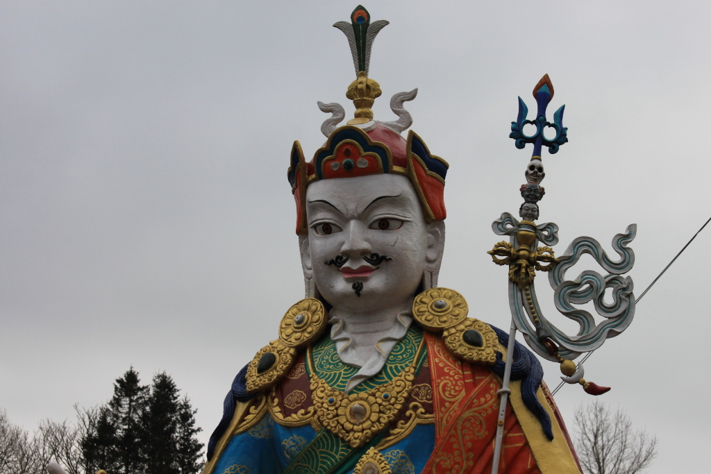 Close-up view of a large, ornate statue, likely of a Buddhist deity.  The statue is predominantly white, with a richly decorated headdress and robes featuring intricate gold and jewel-toned designs in blue, red, and orange. A stylised staff or sceptre with symbolic elements (possibly skulls and a trident) is held by the figure. The background is a muted grey sky, with some dark evergreens visible at the bottom of the frame.