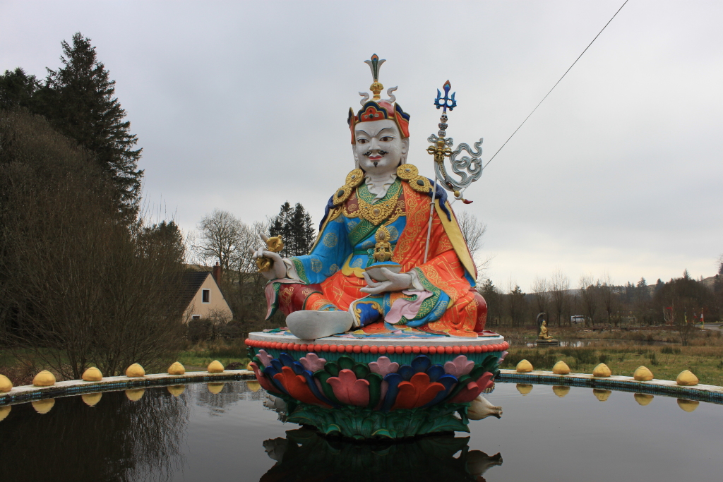 Large, colourful statue of a Buddhist deity, possibly a lama or guru, seated on a lotus throne in a reflecting pool. The statue is richly decorated with intricate details and vibrant colours, including blues, oranges, and golds. The setting appears to be a serene, possibly monastic, landscape with trees and a small building in the background.