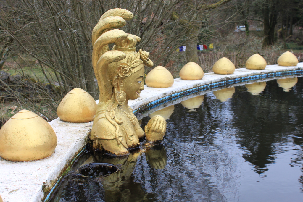 Golden statue of a female figure with multiple cobras forming a crown around her head. The statue is partially submerged in a circular pool of dark water, surrounded by a white border decorated with golden, egg-shaped ornaments. The background features bare winter trees and hints of a garden setting. The statue appears to be in a prayer-like pose.