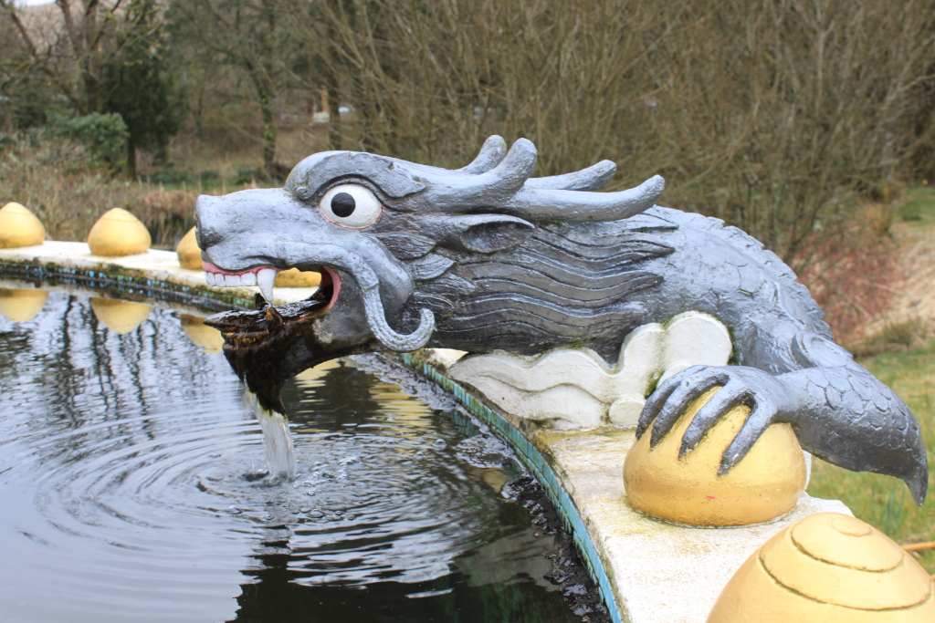 Grey stone dragon statue that is part of a fountain. Water flows from the dragon's mouth into a pool. The dragon rests on a white wall adorned with golden spheres. The background is a blurred-out garden scene with trees and bushes.