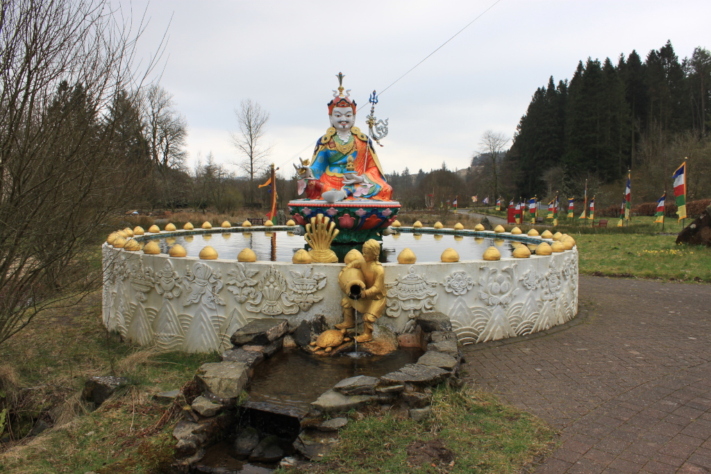 Circular fountain with a statue of a richly adorned Buddhist deity at its centre. The deity is seated and holds a trident-like object. The fountain is ornately decorated with carvings and features small golden spheres around its rim. A smaller golden statue, possibly a yaksha, pours water into the lower part of the fountain.