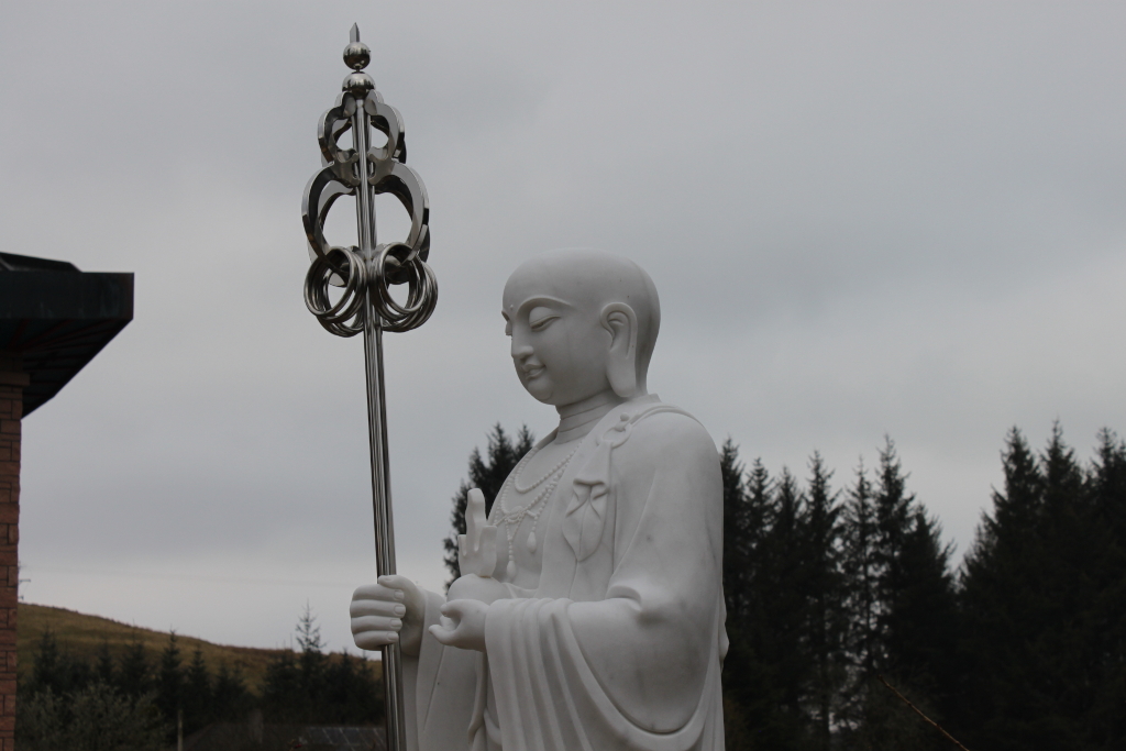 White marble statue of a Buddhist Bodhisattva, holding a silver vajra. The statue is positioned in front of a dark green forest under a gray sky. The overall mood is serene and contemplative. The background is blurred, drawing attention to the statue. A small portion of a building is visible in the lower left corner.