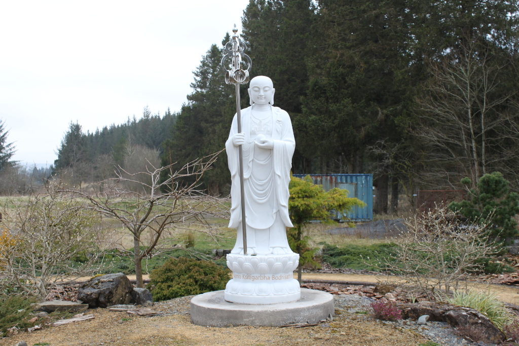 White marble statue of Ksitigarbha Bodhisattva, a Buddhist deity, standing in a garden setting. The statue holds a staff and is positioned on a lotus-shaped base. The background features a coniferous forest and a blue shipping container. The overall impression is one of serenity and spiritual contemplation.