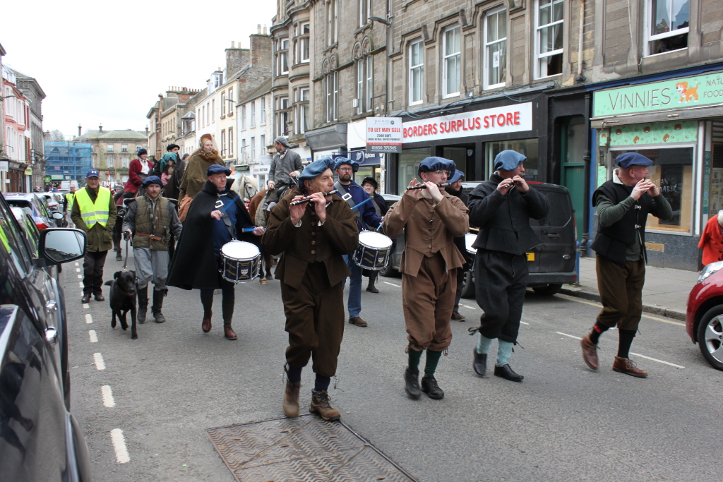 Street parade or procession, possibly a historical reenactment. A group of people dressed in 17th-century style clothing are marching down a street lined with buildings. Some are playing musical instruments (flutes and drums), while others are walking alongside or riding horses. A dog is walking with the group. The overall atmosphere suggests a community event or a celebration.