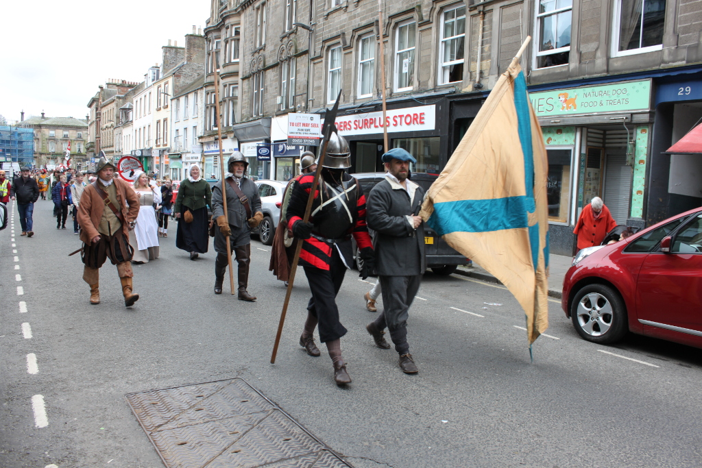 Historical reenactment or parade taking place on a street lined with shops. A group of people are dressed in 17th-century-style clothing, some in military attire, marching down the street. They appear to carrying a large banner or flag with a teal cross on a beige background. Spectators and modern-day vehicles are visible in the background, creating a juxtaposition between the historical reenactment and contemporary life. The overall tone is one of a community event or historical commemoration.