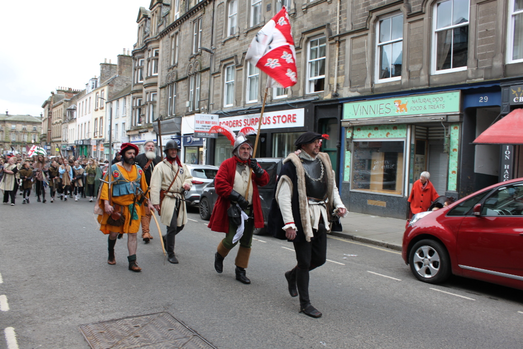 Procession of people in period costumes, likely participating in a historical reenactment or parade. The costumes appear to be from the late medieval or early Renaissance periods, with several individuals carrying weaponry (long spears or halberds are visible). A prominent red and white flag is carried at the front of the procession. The event is taking place on a street in a town, with shops visible in the background. The overall impression is one of historical recreation, possibly celebrating a specific historical event or period. The emotional tone is one of festive participation and historical engagement. The moral of the image, if any, is implicitly about the appreciation and commemoration of history.