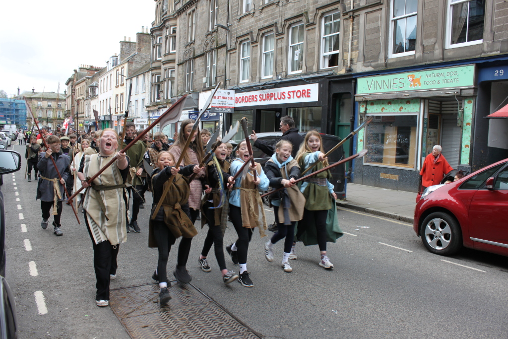 Group of children dressed in medieval-style costumes, marching down a street. They are carrying wooden spears or staffs, and some are shouting or making excited expressions. The setting appears to be a town or city in the UK, judging by building styles and signage. The children are the central focus, creating a sense of movement and energy within the overall scene. The background includes shops and onlookers, suggesting a parade or historical reenactment.