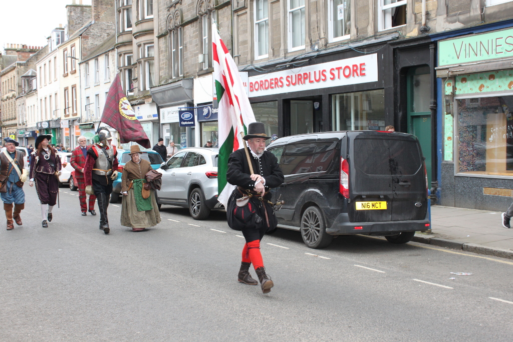 Parade or procession of people dressed in 17th-century style clothing, walking down a street lined with shops. A man carrying a flag is prominently featured in the centre. The overall scene suggests a historical re-enactment or a celebratory event.
