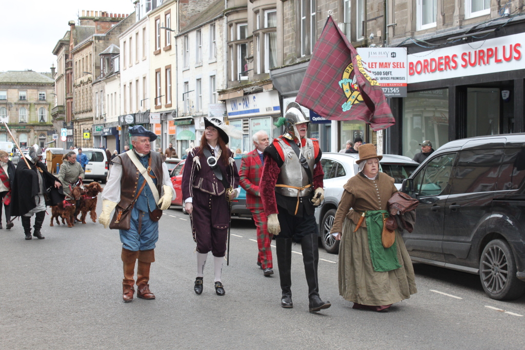 Procession of people in 17th-century-style clothing walking down a street. There are men and women in period costumes, some appearing as military figures, others in civilian attire. Two dogs are also walking with the group. The setting is a modern town with shops and cars visible in the background, suggesting a historical re-enactment or parade. A tartan flag is prominently carried.