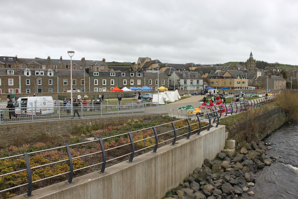 Riverside scene in a Hawick, Scottish Borders within in the UK. A community event or market is taking place in a paved area, with numerous people milling about and several colourful marquees or tents set up. The background consists of rows of traditional stone buildings. A low concrete wall with a metal railing separates the event area from a riverbank, which is reinforced with rocks. The overall mood appears to be one of a typical, perhaps slightly overcast, day in a small town.