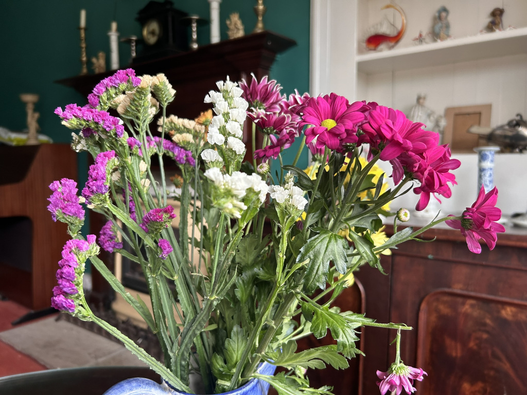 Bouquet of flowers in a blue vase. The bouquet is composed of various flowers including vibrant pink chrysanthemums, smaller purple statice, and white gypsophila. The bouquet sits on a table in front of a dark wood cabinet and a fireplace mantel, with various decorative items visible on the mantel and in the background. The overall setting appears to be in a home, perhaps a living room, with a slightly vintage or antique aesthetic.