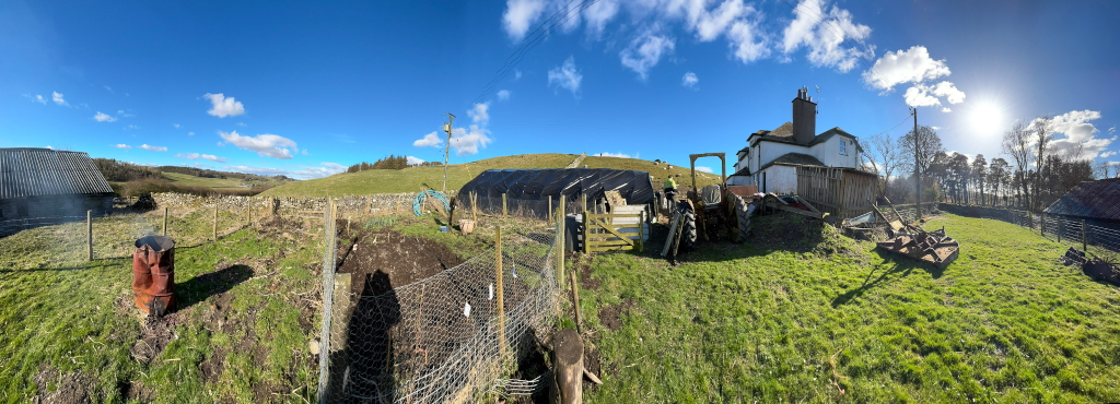 A panoramic view of a rural property on a sunny day. The foreground shows a small garden area with wire fencing, a compost heap, and an old rusty barrel. A tractor is parked near a farmhouse, which is a white building with a dark roof. In the background, there are rolling hills, some trees, and other farm buildings. The overall scene suggests a working farm or smallholding, possibly under development given the tractor and what appears to be a newly constructed or prepared garden plot. The sun is bright, creating shadows that define the landscape.