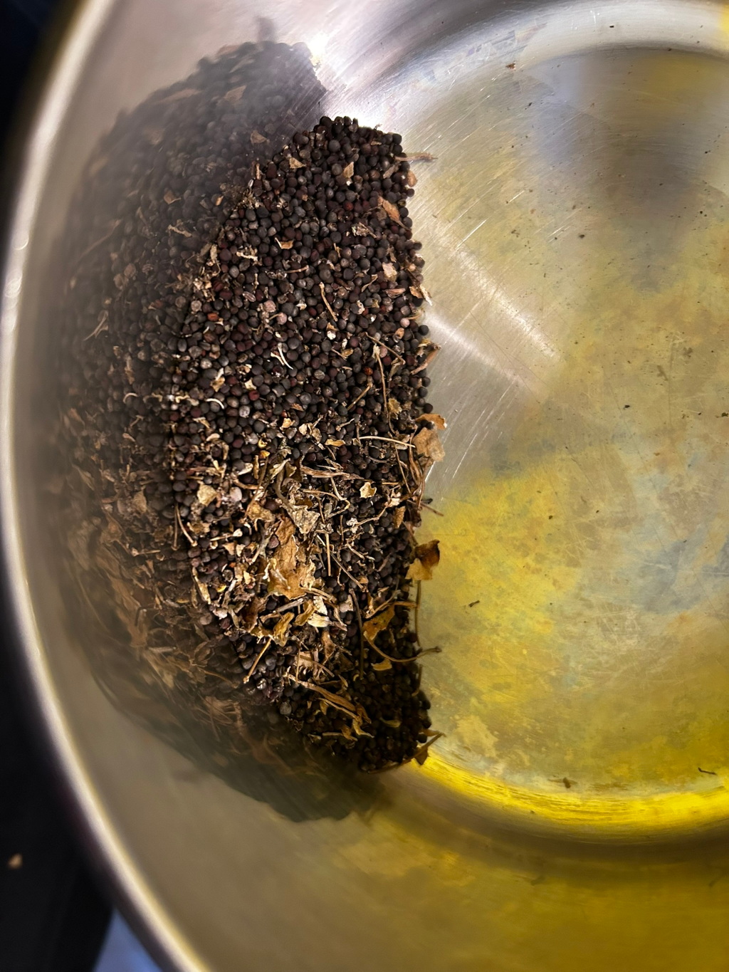 Metal bowl containing a mixture of dark-coloured mustard seeds and dried herbs or spices. The mixture is piled in one side of the bowl, leaving the other side largely empty. The bowl appears to be metallic and shows signs of wear or discolouration.