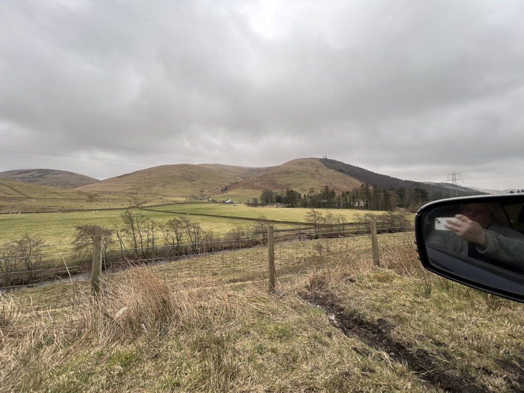 A pastoral landscape under a cloudy sky. Leonie is taking a selfie in a car's side mirror, with the rolling hills and a fence in the background. The scene is peaceful and somewhat muted in color due to the overcast weather. The overall impression is one of tranquil countryside.