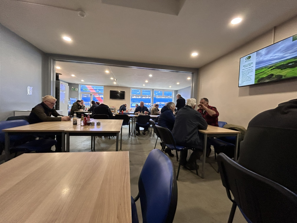 Room with several tables and chairs, where a group of people are seated, seemingly engaged in conversation. The room appears to be a cafeteria with simple light wood furniture. A large window looks out onto an area.