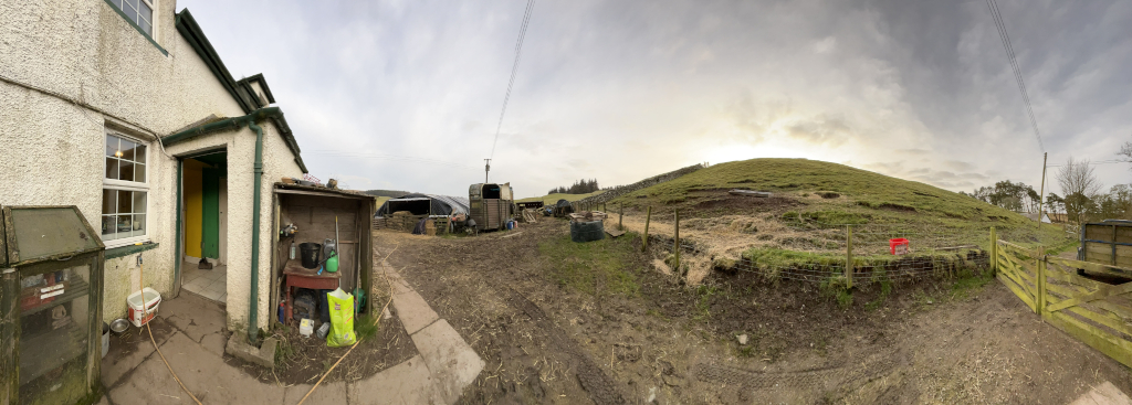 Panoramic view of a rural farmstead. The foreground features a section of a whitewashed stone building with a green door, and various farm implements and storage units. The mid-ground shows a muddy farmyard with a horse trailer, livestock shelters, and stacks of hay. In the background, a sloping grassy hill rises gently. The overall impression is one of a working farm, possibly in a remote location. The sky is overcast and slightly grey.