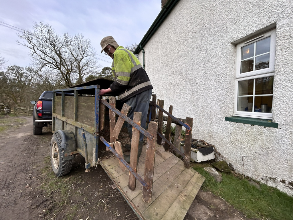 Charlie in a high-visibility vest unloading something from a small livestock trailer attached to a pickup truck parked next to a whitewashed building. The trailer's ramp is down, and Charlie appears to be carefully handling the contents.