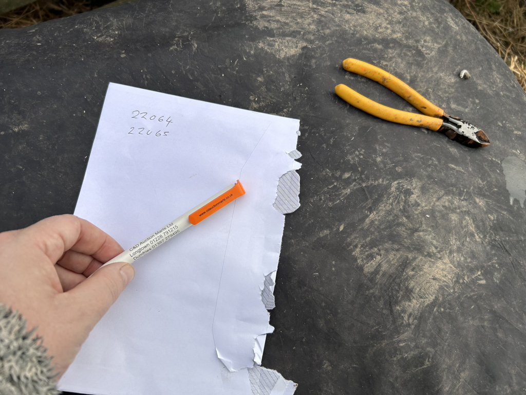 A piece of white paper with the numbers 22064 and 22065 written on it. A pen with the logo of C&D Auction Marts Ltd is resting on the paper, and a pair of yellow wire cutters is lying on a dark-coloured surface nearby. The paper appears to have been torn. The overall context suggests some kind of field work or inventory taking, possibly related to the auction house.