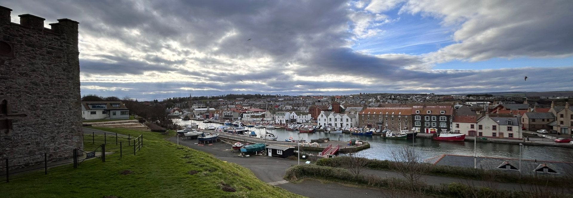 Panoramic view of a harbour town under a cloudy sky. A stone tower is prominent in the foreground, with a grassy area leading down to a harbour filled with various boats and ships. The town's buildings, many appearing to be multi-story, line the harbour.