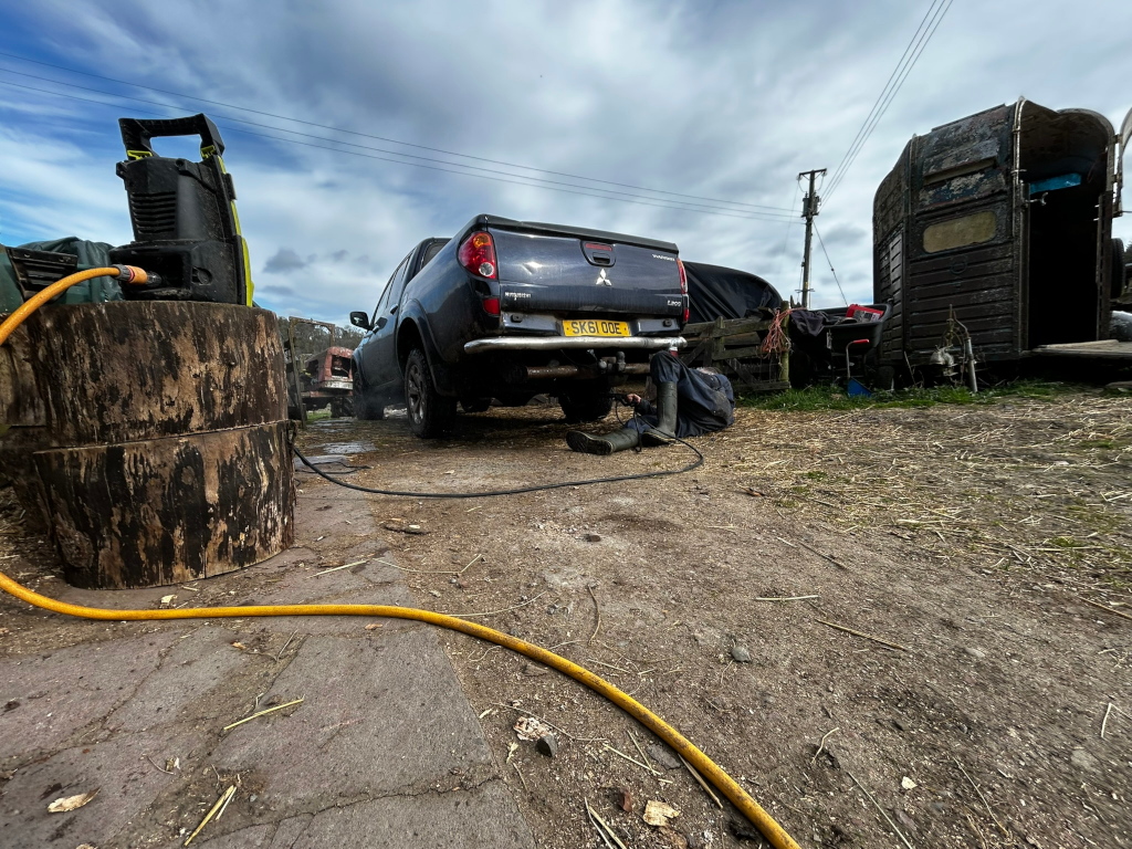 Charlie lying under a dark-grey Mitsubishi L200 pickup truck, cleaning mud from the trucks chassis. A pressure washer is visible in the foreground, along with a yellow hose. The setting is in a rural area, by a farm house, with a horse trailer visible in the background. The overall impression is one of everyday work or maintenance in a rustic setting.