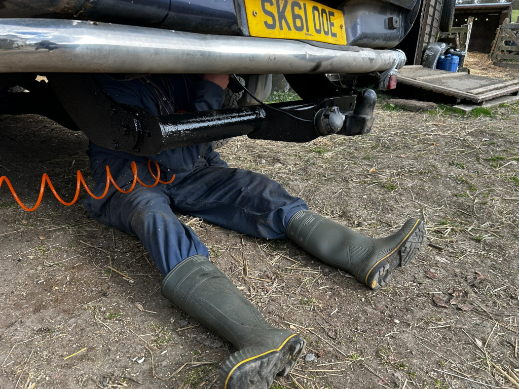 Charlie in blue overalls and green Wellington boots lying under the rear of his blue L200 pick up truck. He is spraying underneath near his trucks tow hitch. An orange coiled air hose is visible near his legs.
